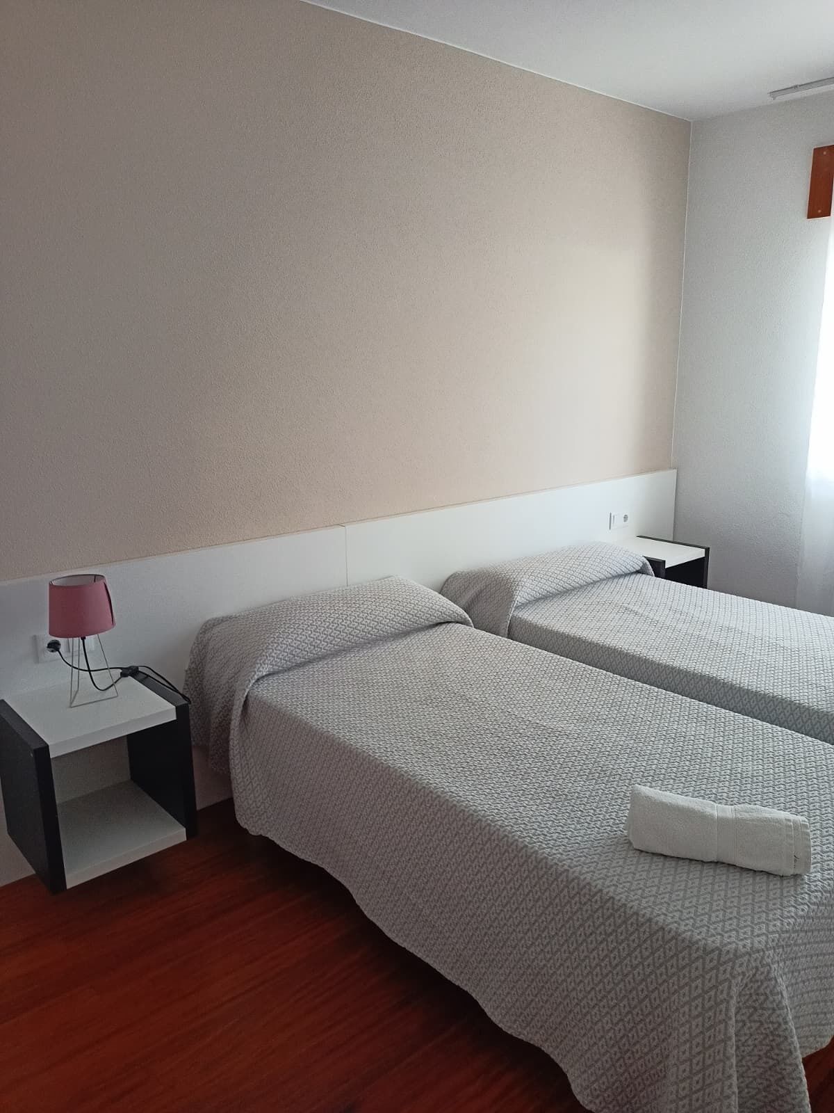 A hotel room with two beds, a bedside table with a lamp, and a wooden floor.