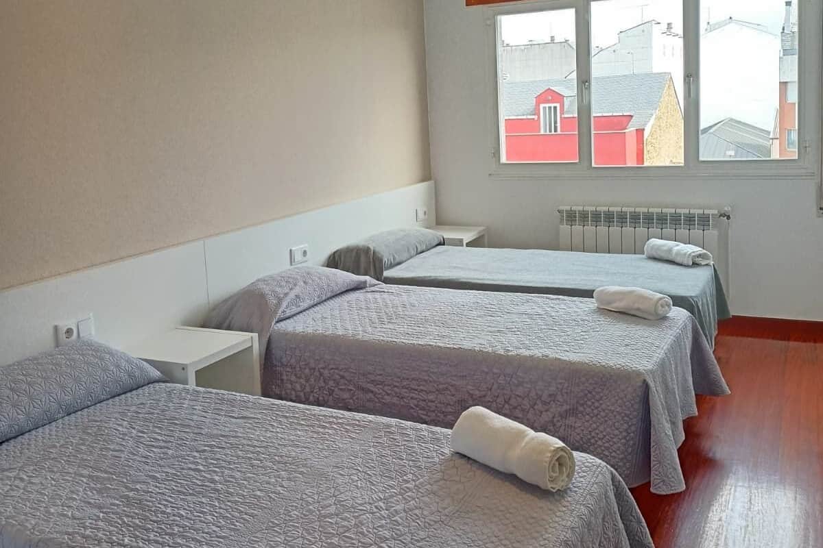 A hotel room with three beds, a window with a view of buildings, a radiator, and a ceiling light.