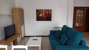 A modern living room with a teal sofa, a white coffee table, a TV on a wooden stand, a painting on the wall, and a wooden cabinet.