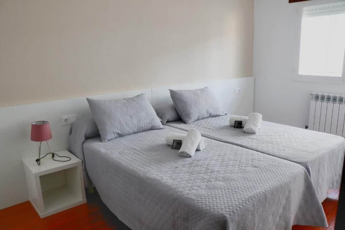 A hotel room with two beds, a bedside table with a lamp, and a window with blinds.