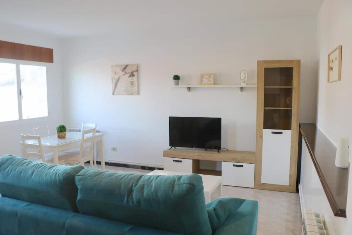 A living room with a teal sofa, a TV on a wooden stand, a dining table with chairs, and a small plant on the wall.