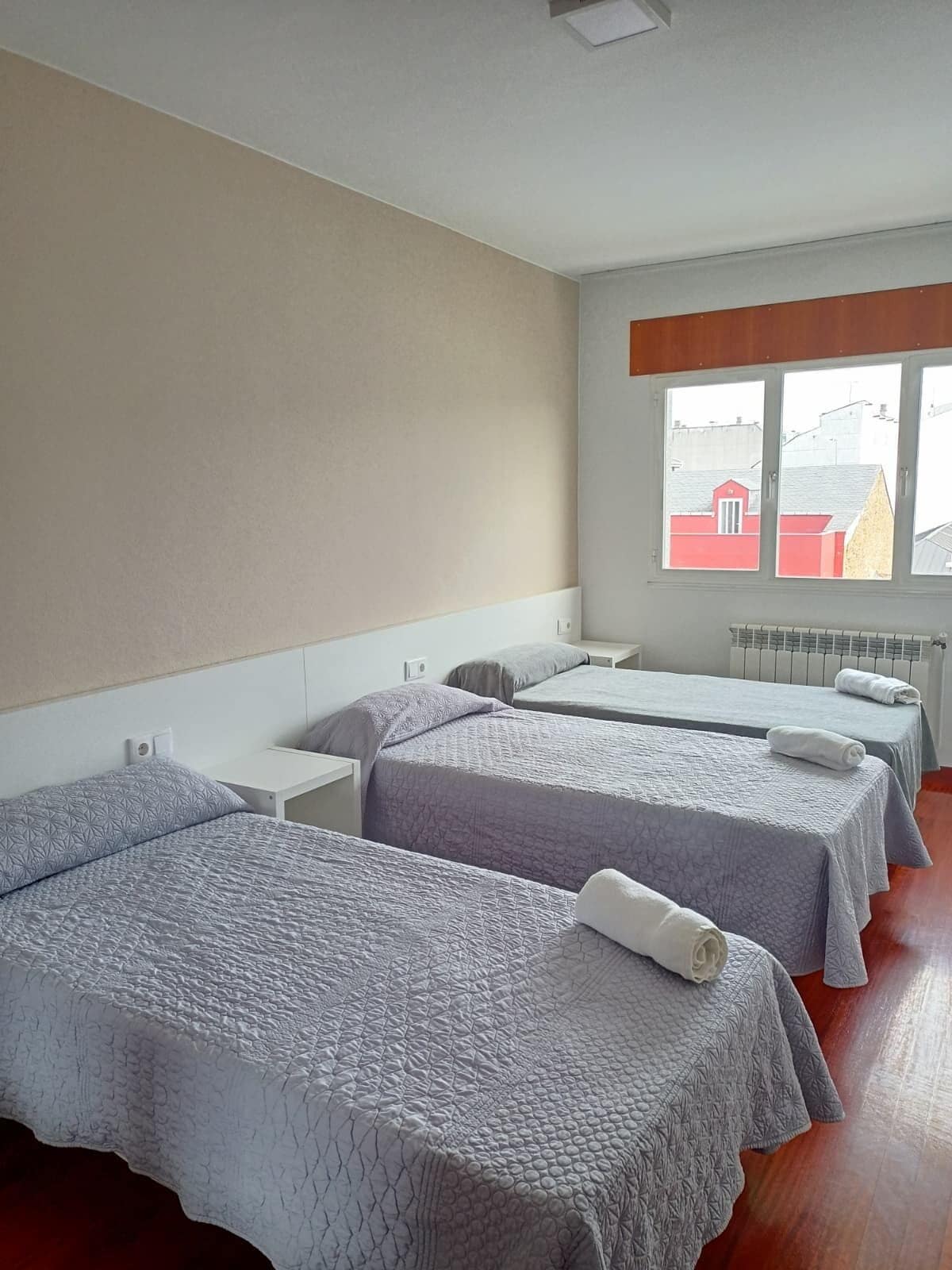 Une chambre d’hôtel avec trois lits, une fenêtre donnant sur des immeubles et un radiateur.