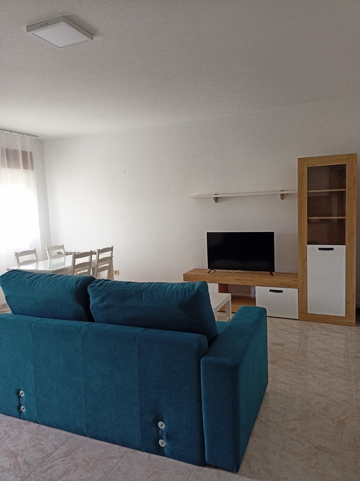 Un salon avec un canapé de couleur turquoise, un téléviseur posé sur un meuble en bois, une table à manger avec des chaises et un buffet aux portes vitrées.
