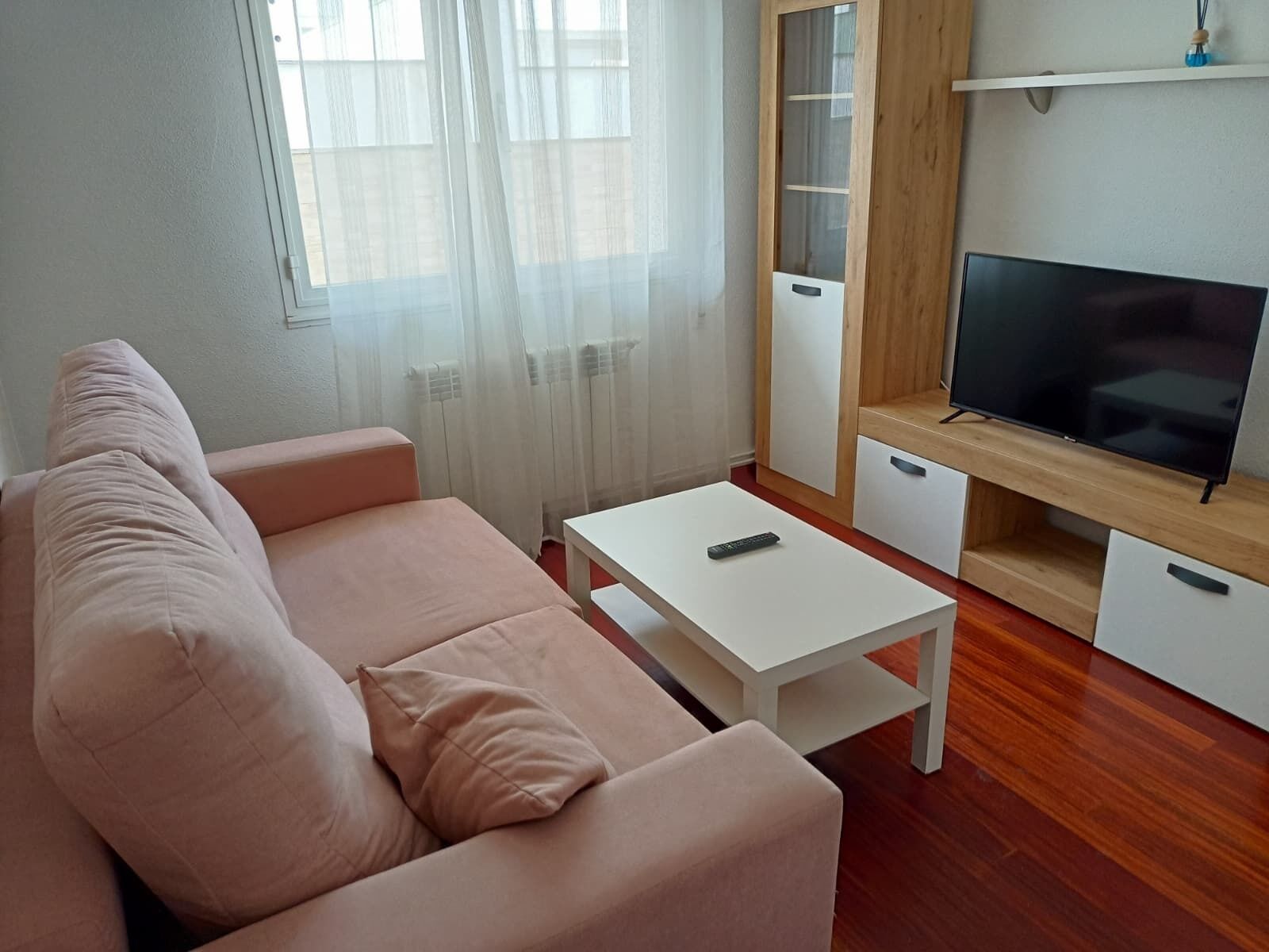 A living room with a sofa, a coffee table, a TV on a wooden stand, and a window with sheer curtains.