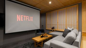 Un salon moderne doté d’un grand écran affichant le logo « NETFLIX », d’une table basse en bois, d’un canapé gris et d’une porte coulissante de style japonais traditionnel.