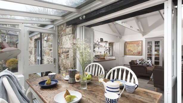 A dining area with a wooden table, white chairs, and a stone wall. - Rosehendra - A beautifully refurbished pet-friendly barn conversion with two bedrooms, a log burner (Rosevidney)