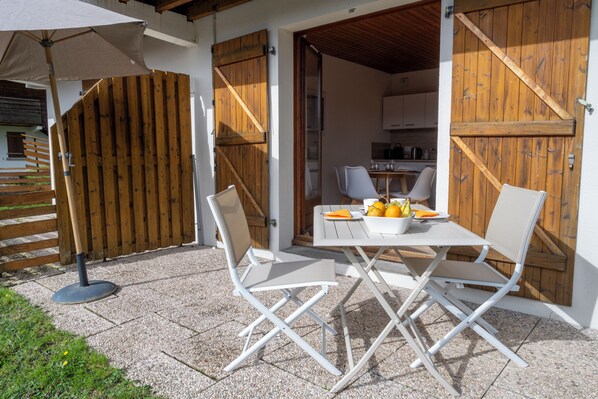 Um pátio externo com mesa e cadeiras, cerca de madeira e cozinha visível por uma porta aberta.