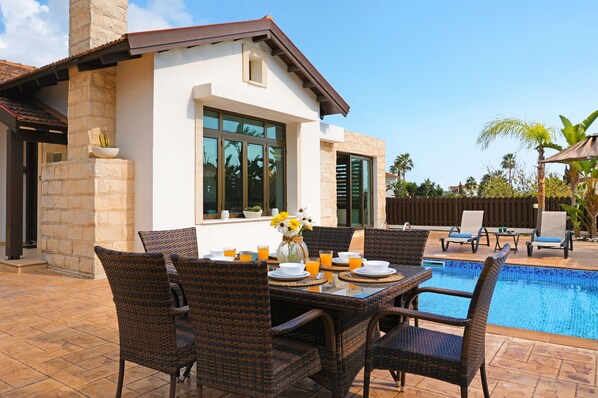 Un patio con mobiliario de mimbre, una mesa de comedor con platos y vasos, y una piscina.