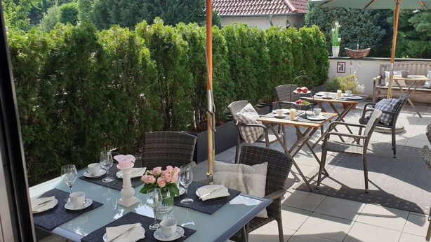 En udendørs patio med borde dækket til spisning, omgivet af grøn vegetation og et flisebelagt gulv.