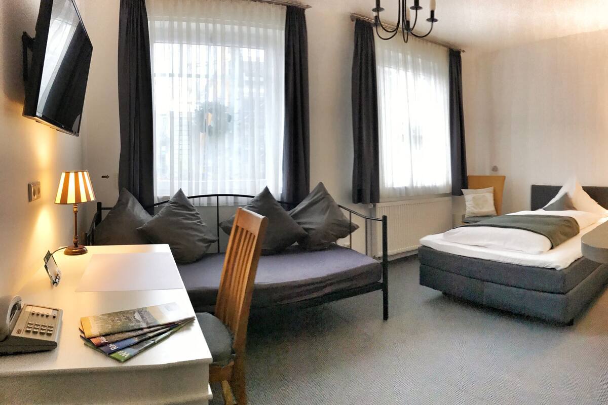 Hotellihuone, jossa on sänky, kirjoituspöytä lampun kanssa, tuoli ja pieni keittonurkkaus.