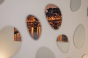 A view through a patterned wall with circular cutouts, revealing a nighttime cityscape with illuminated buildings and a bridge over water.