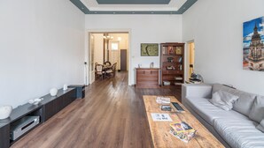 Un salon moderne avec un plancher en bois, un canapé gris, une table basse en bois sur laquelle sont posés des magazines, un meuble pour la télévision et un coin repas agrémenté d’un lustre.