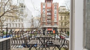 Une vue depuis un balcon donnant sur une cour intérieure, avec une table et des chaises en bois, un bâtiment rouge en arrière-plan et des voitures garées.
