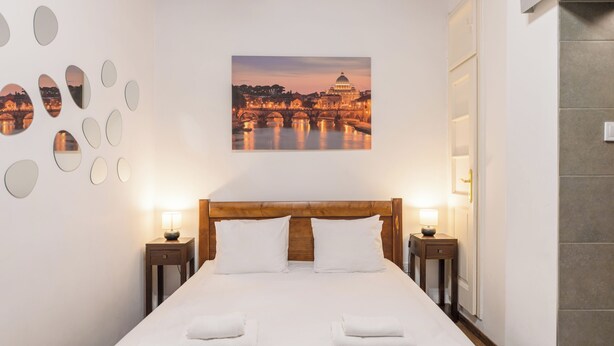 Une chambre d’hôtel avec un grand lit, deux tables de chevet avec des lampes, un tableau représentant un paysage urbain accroché au mur, et une série de miroirs ronds disposés sur le mur.