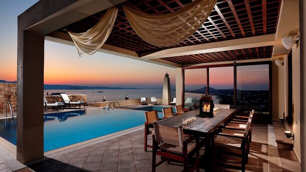 Een dakzwembad met uitzicht op een zonsondergang, een eettafel met stoelen, en een pergola met draperende stof.