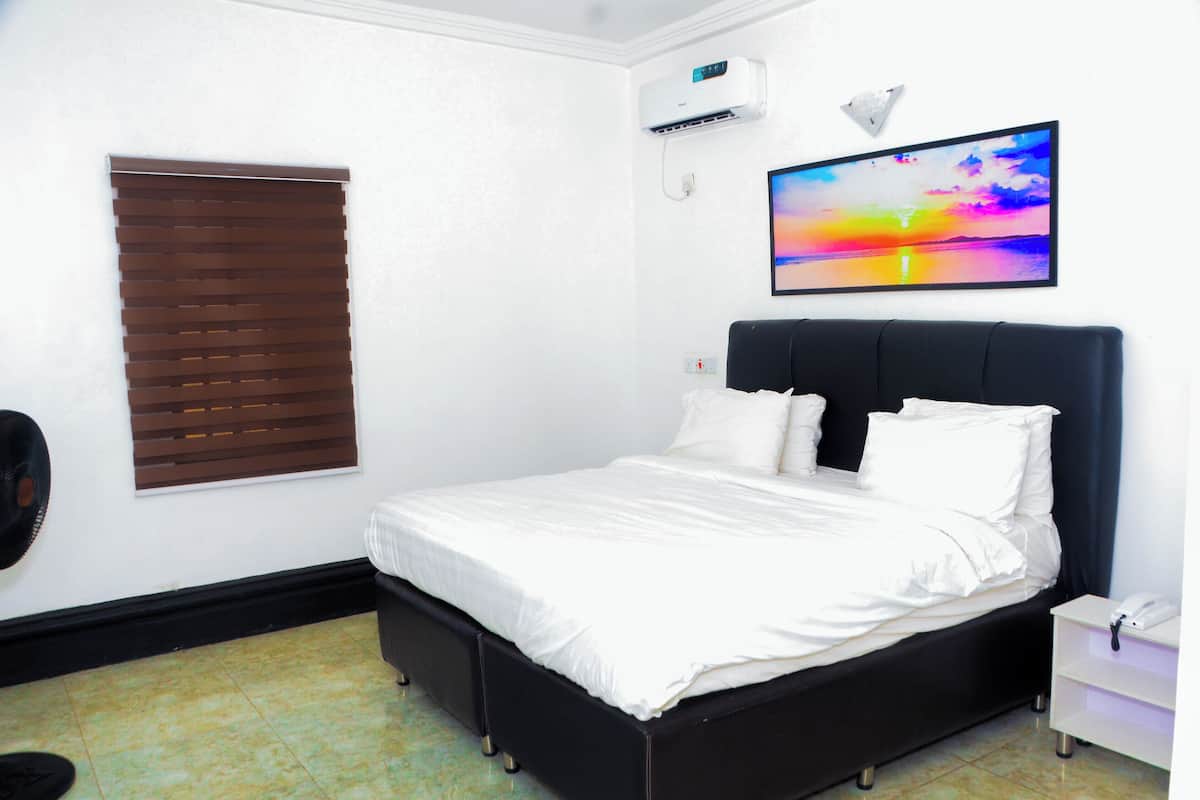 A bedroom with a bed, a wall-mounted air conditioner, a window with blinds, and a TV on the wall.