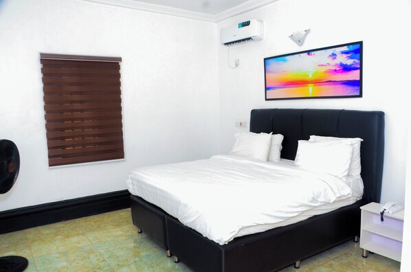 A bedroom with a bed, a wall-mounted air conditioner, a window with blinds, and a TV on the wall.