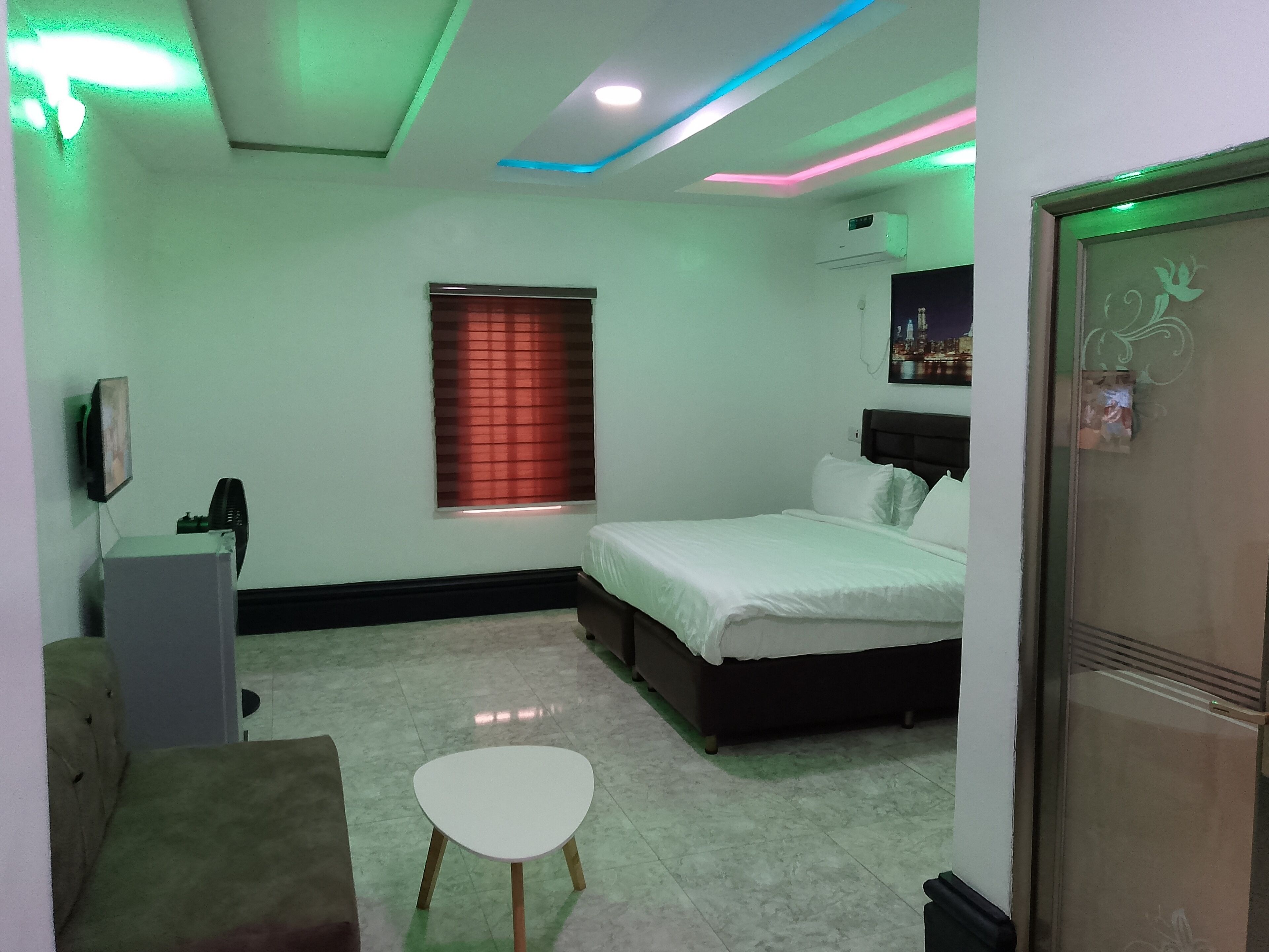 A bedroom with a bed, a desk, a chair, a window with blinds, and an air conditioning unit.