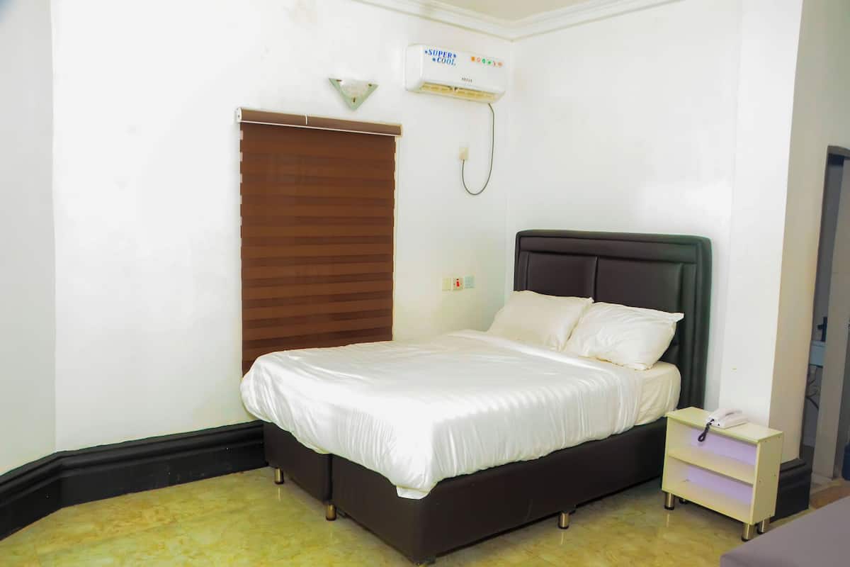 A neatly made bed with white linens in a room with a brown headboard, a small bedside table with a phone, and an air conditioning unit on the wall.