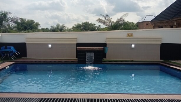 A swimming pool with a waterfall feature, surrounded by a low wall and a covered area with blue roofing. - FMMM1 (Agbor)