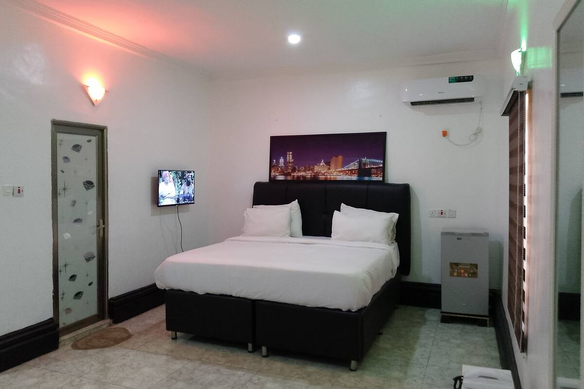 A hotel room with a bed, a television, a nightstand, and a wall-mounted air conditioner.