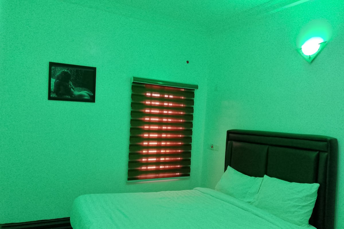 A room with a bed, a window with blinds, a wall-mounted light, and a small picture on the wall.