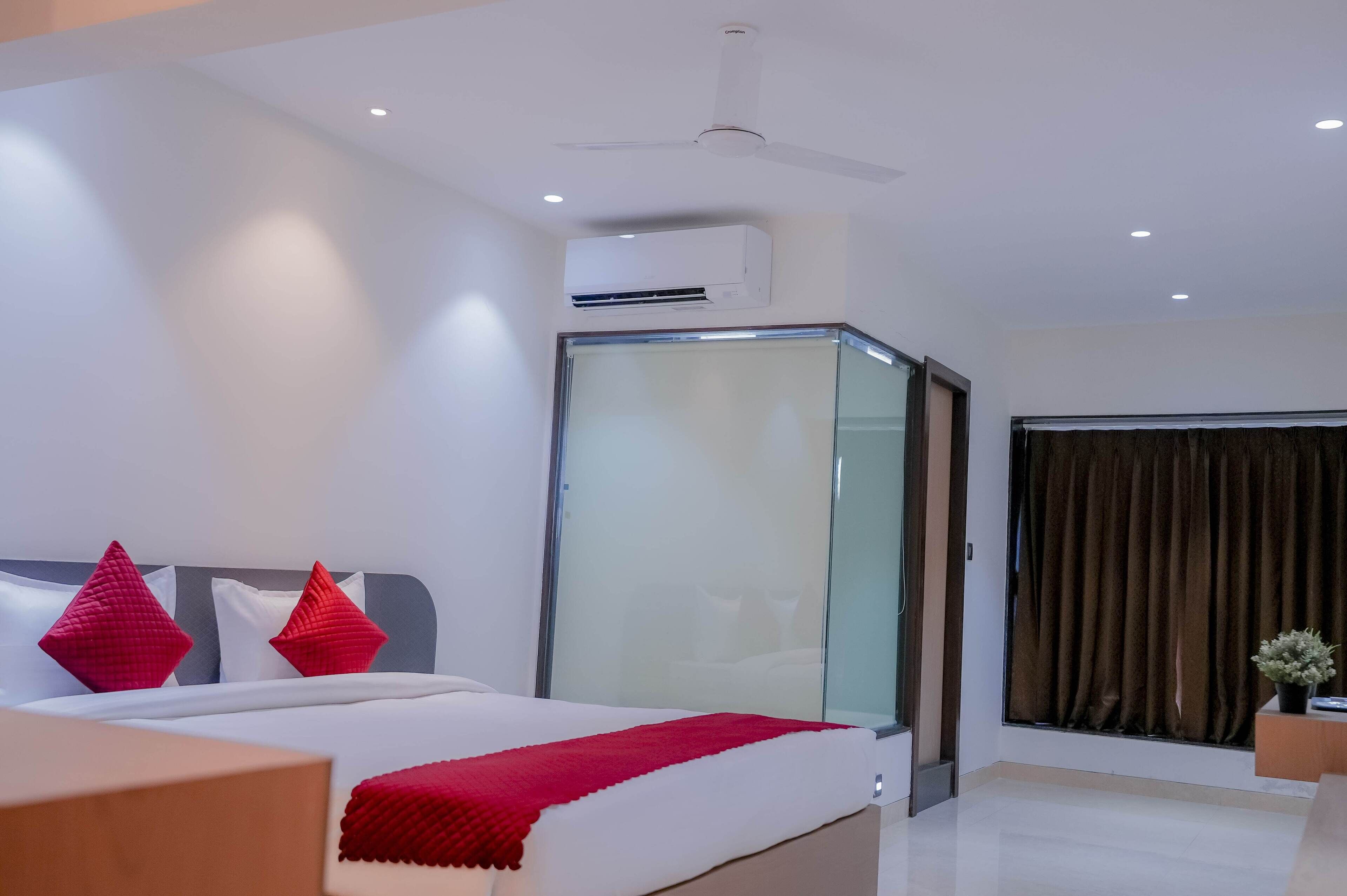 A modern hotel room with a bed, red pillows, a glass partition, a ceiling fan, an air conditioning unit, and a window with curtains.