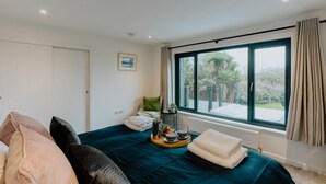 A bedroom with a large bed, a view of the outdoors, a window with curtains, and a bedside table with a tray of breakfast items.