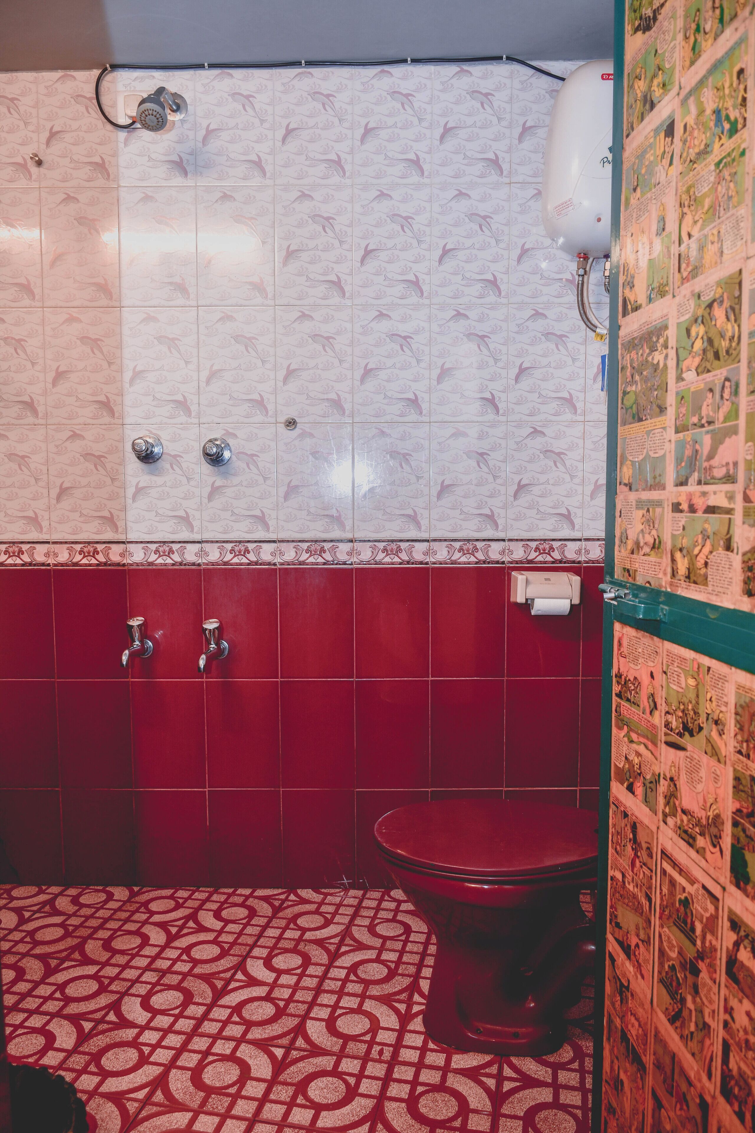 A bathroom with a red and white tiled floor, a toilet, a showerhead, and a wall with comic strip decorations.