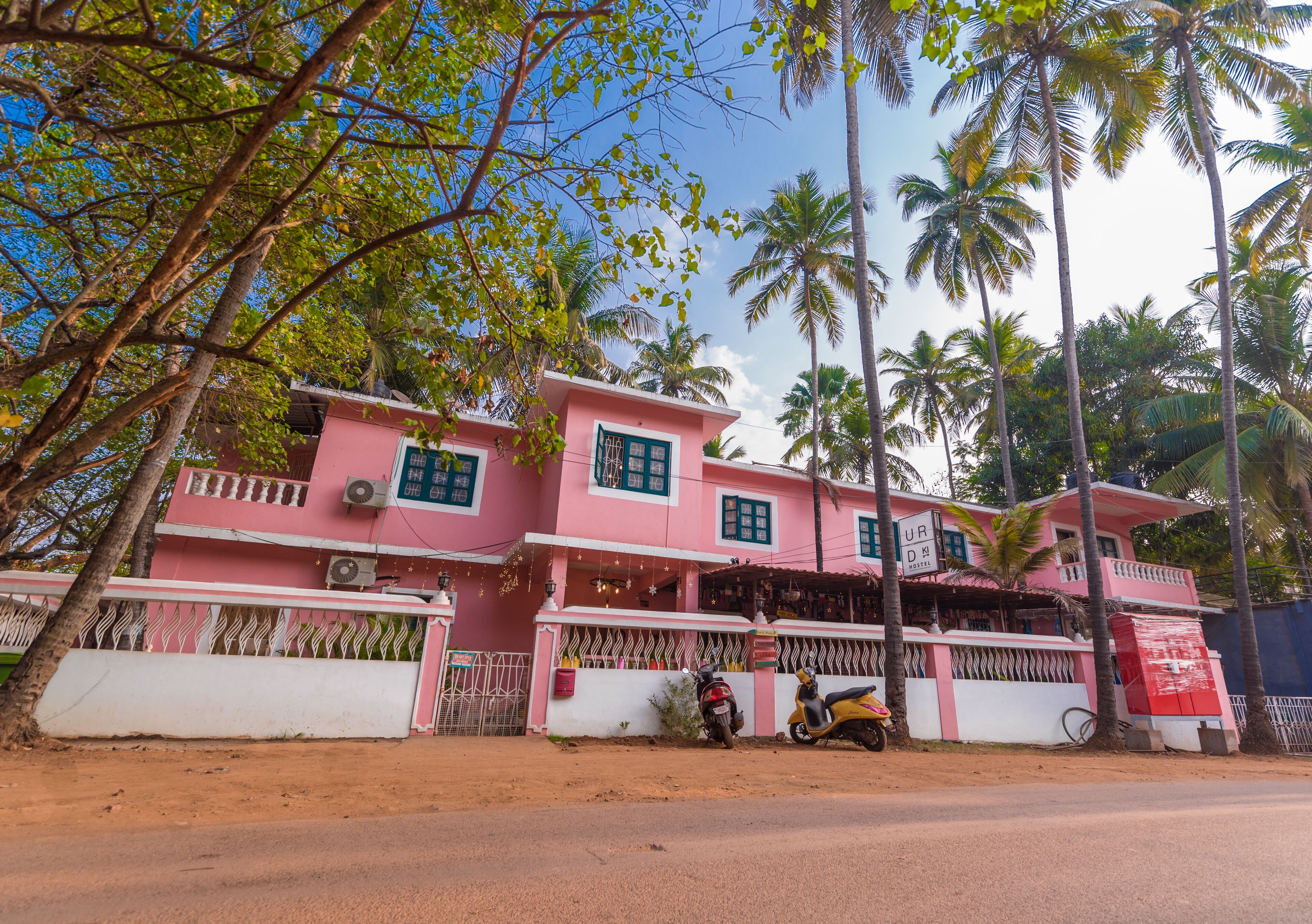 A pink two-story building with a gated entrance, a parked scooter, and palm trees in the background.