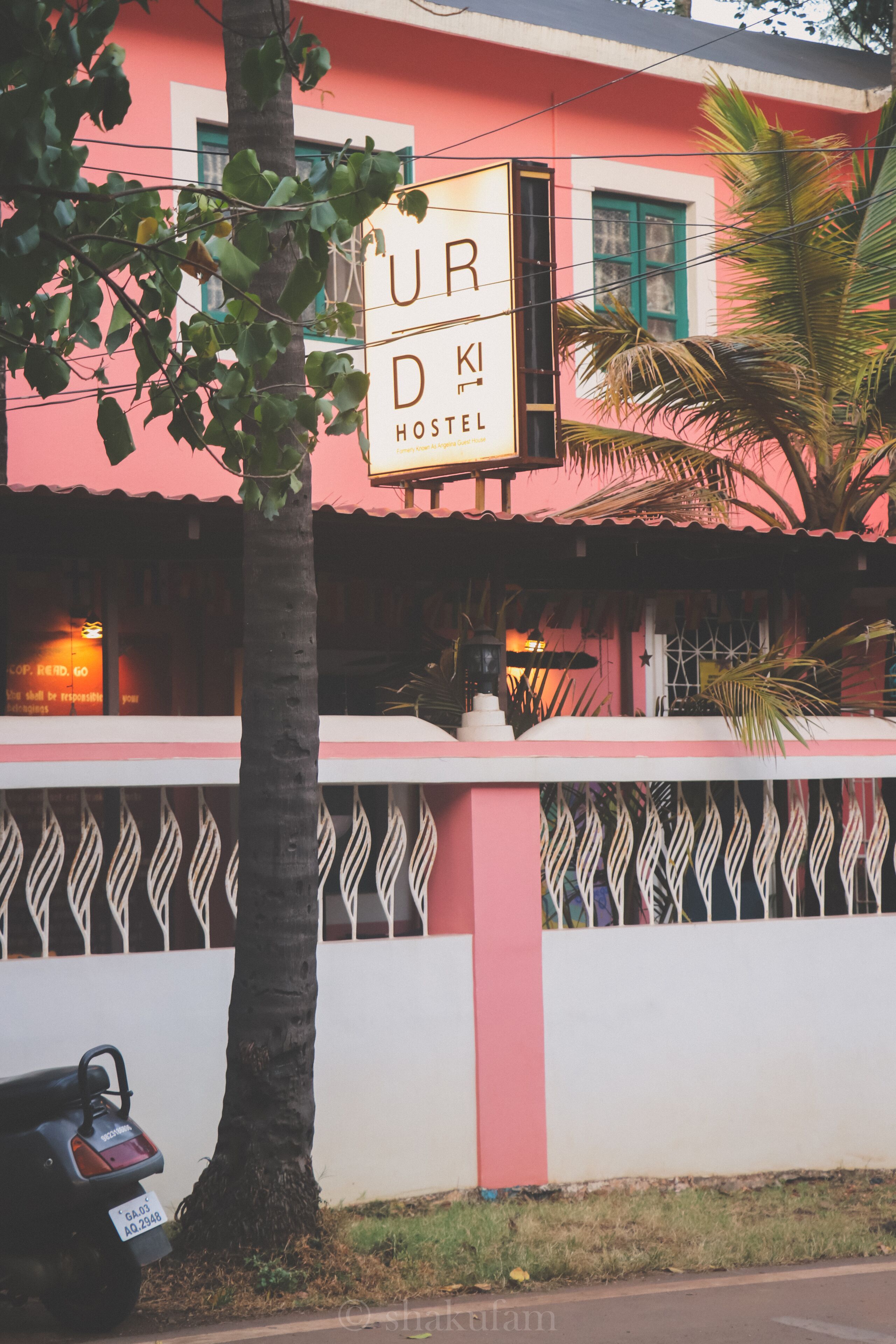 A hostel with a pink facade, a signboard, and a tree in front.