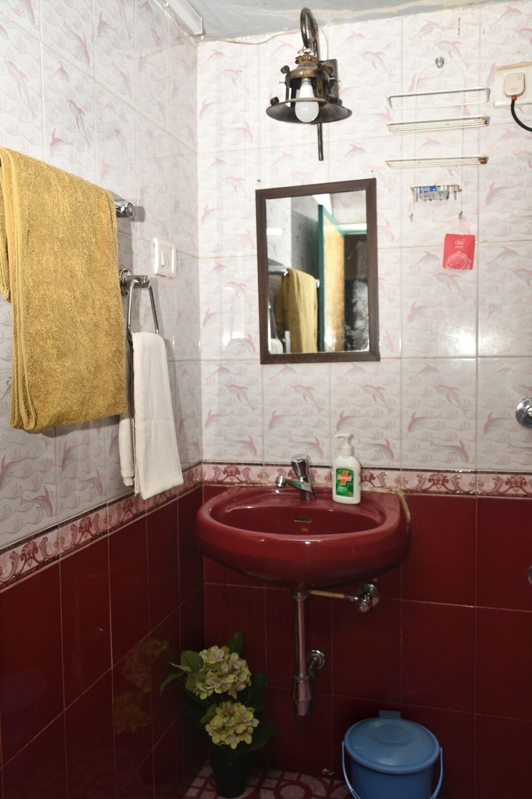 A bathroom with a red sink, a mirror, a towel rack with a gold towel, a bottle of hand soap, and a blue trash bin.