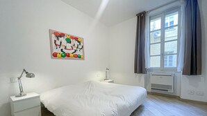 A bedroom with a bed, two bedside tables, a desk lamp, a wall-mounted air conditioner, and a window with curtains.