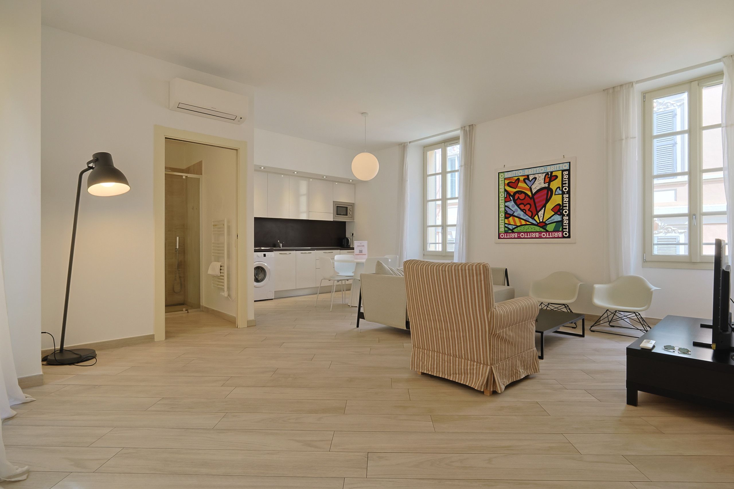 A modern living room with a sofa, a coffee table, and a TV. There is a kitchen area with a microwave and a washing machine. The room has large windows and a ceiling light.
