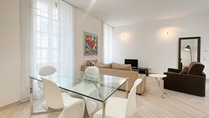 A modern living room with a glass dining table, white chairs, a beige sofa, and a brown armchair.