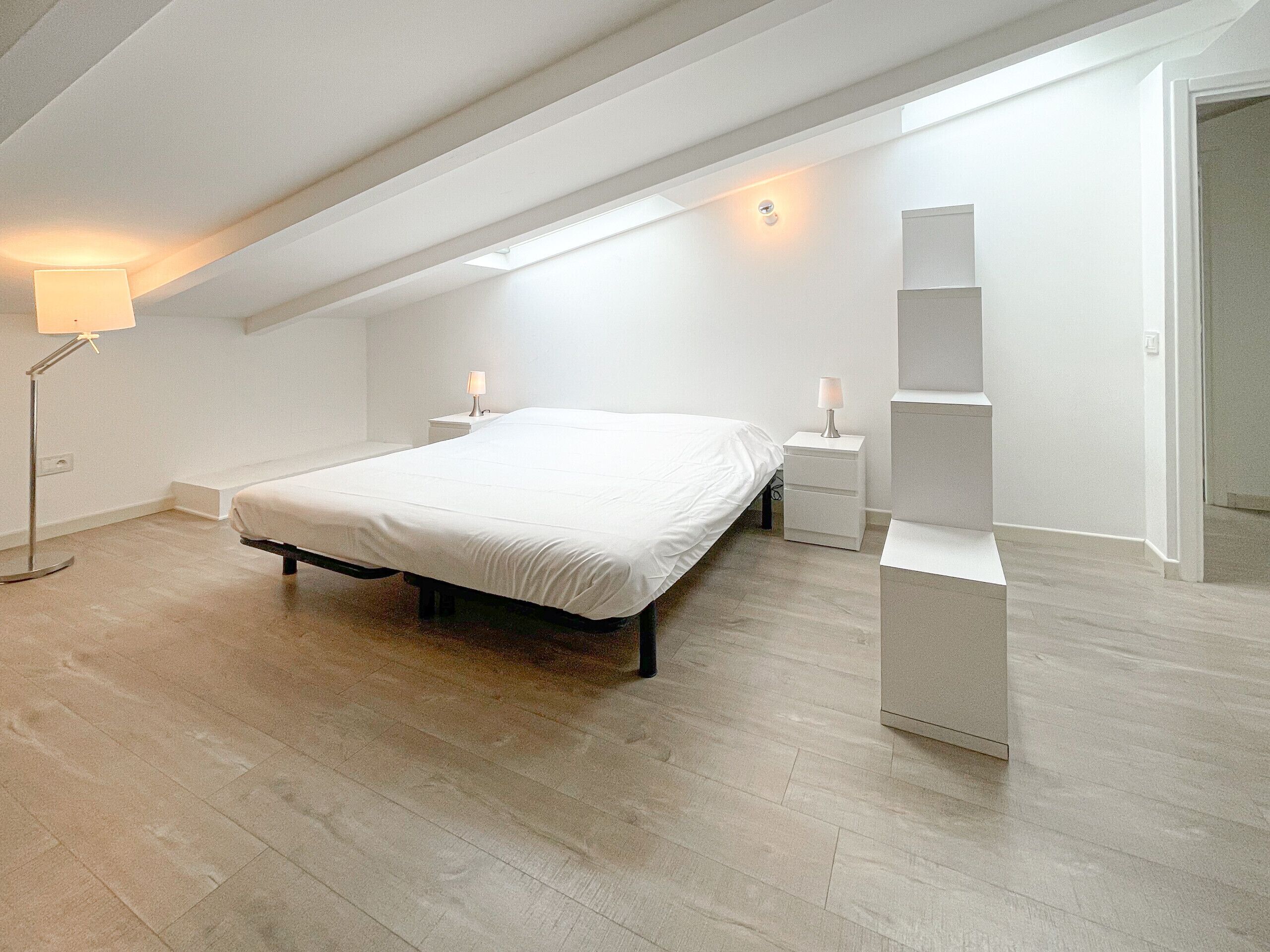 Et moderne, minimalistisk soveværelse med en stor seng, sengeborde med lamper og et indbygget reolsystem.