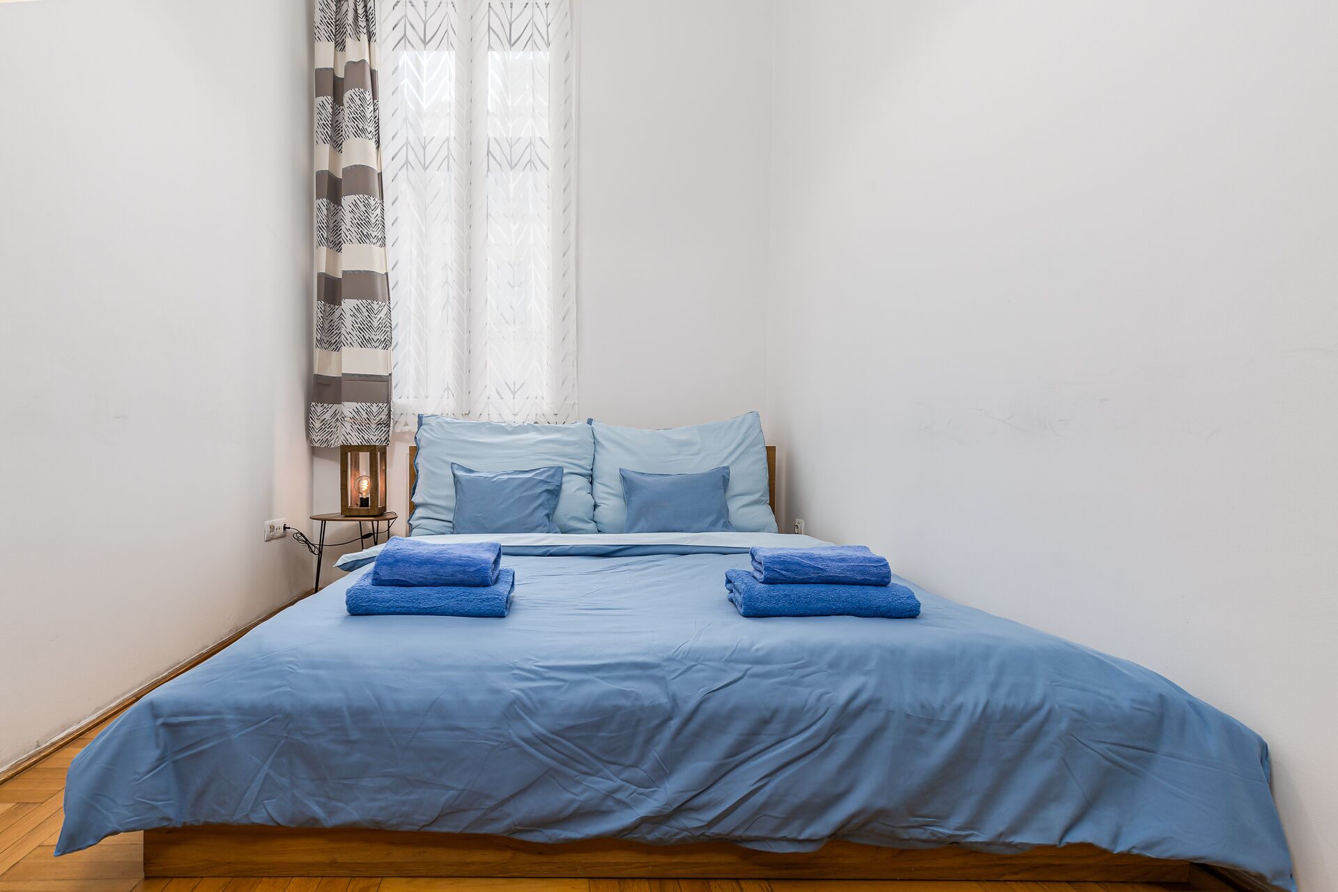 Una cama bien tendida con ropa de cama y almohadas azules, una mesita de noche con una lámpara y cortinas estampadas.