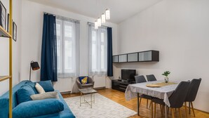 A modern living room with a blue sofa, a dining table with chairs, a TV, and a radiator. - Venus Apartments - Gozsdu Court III (Budapest)