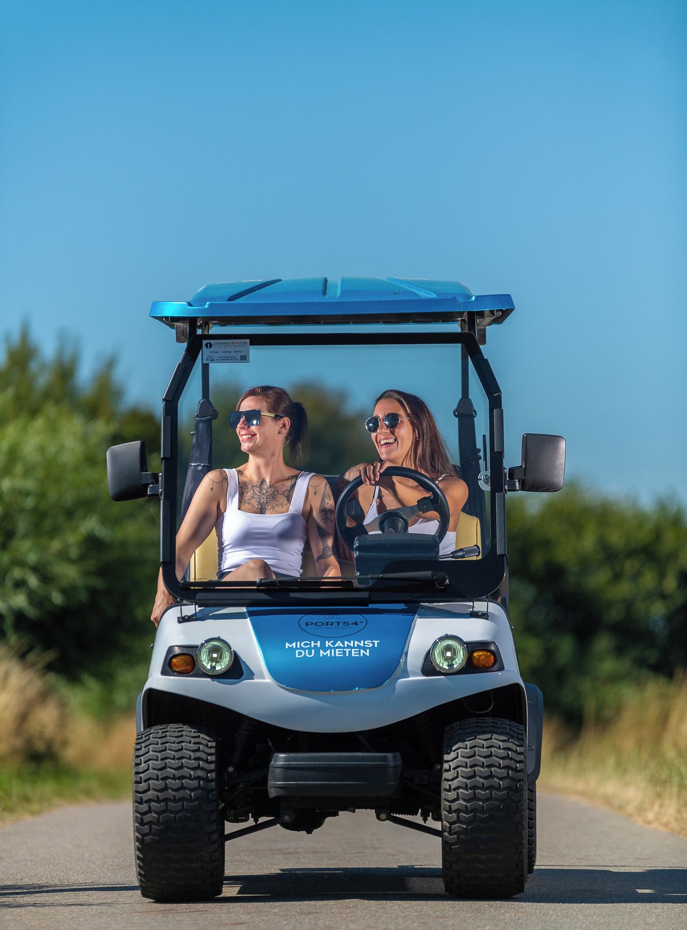 Une voiturette de golf avec deux personnes à bord, dotée d’un toit bleu et de l’inscription « MICH KANNST DU MIETEN ».