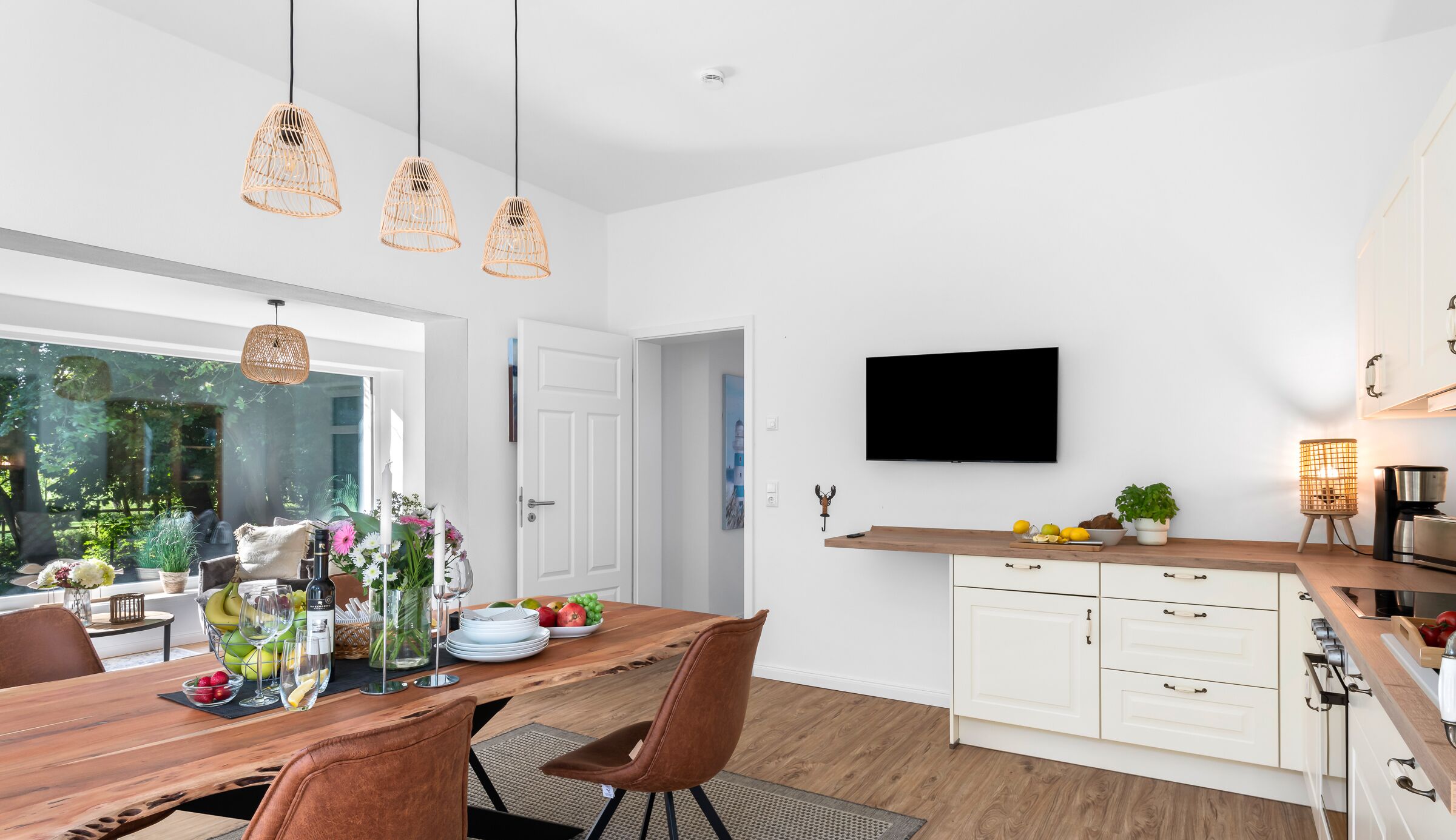 Una cocina moderna con armarios blancos, encimera de madera y estufa integrada. Hay un comedor con mesa y sillas de madera, y un ventanal con vistas al verdor.