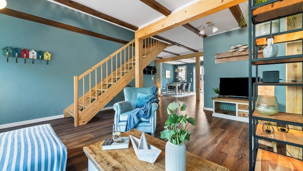 Un salón amplio con una escalera de madera, un sillón azul y una mesa de centro de madera con un jarrón de flores.