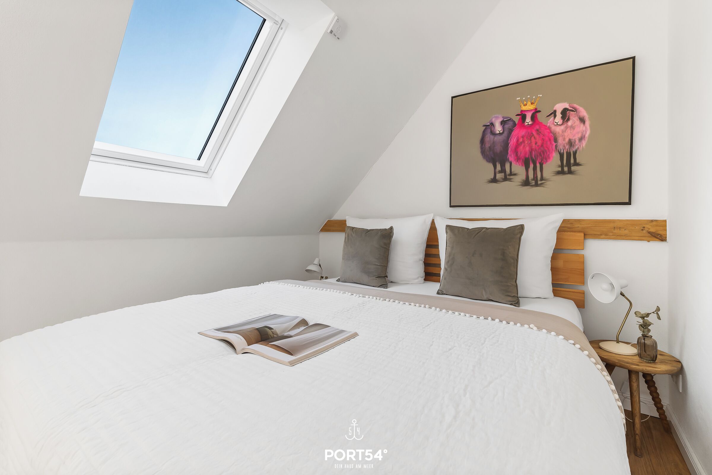 Een slaapkamer met een schuine plafond, een groot bed, een nachtkastje met een lamp, en een schilderij van schapen aan de muur.