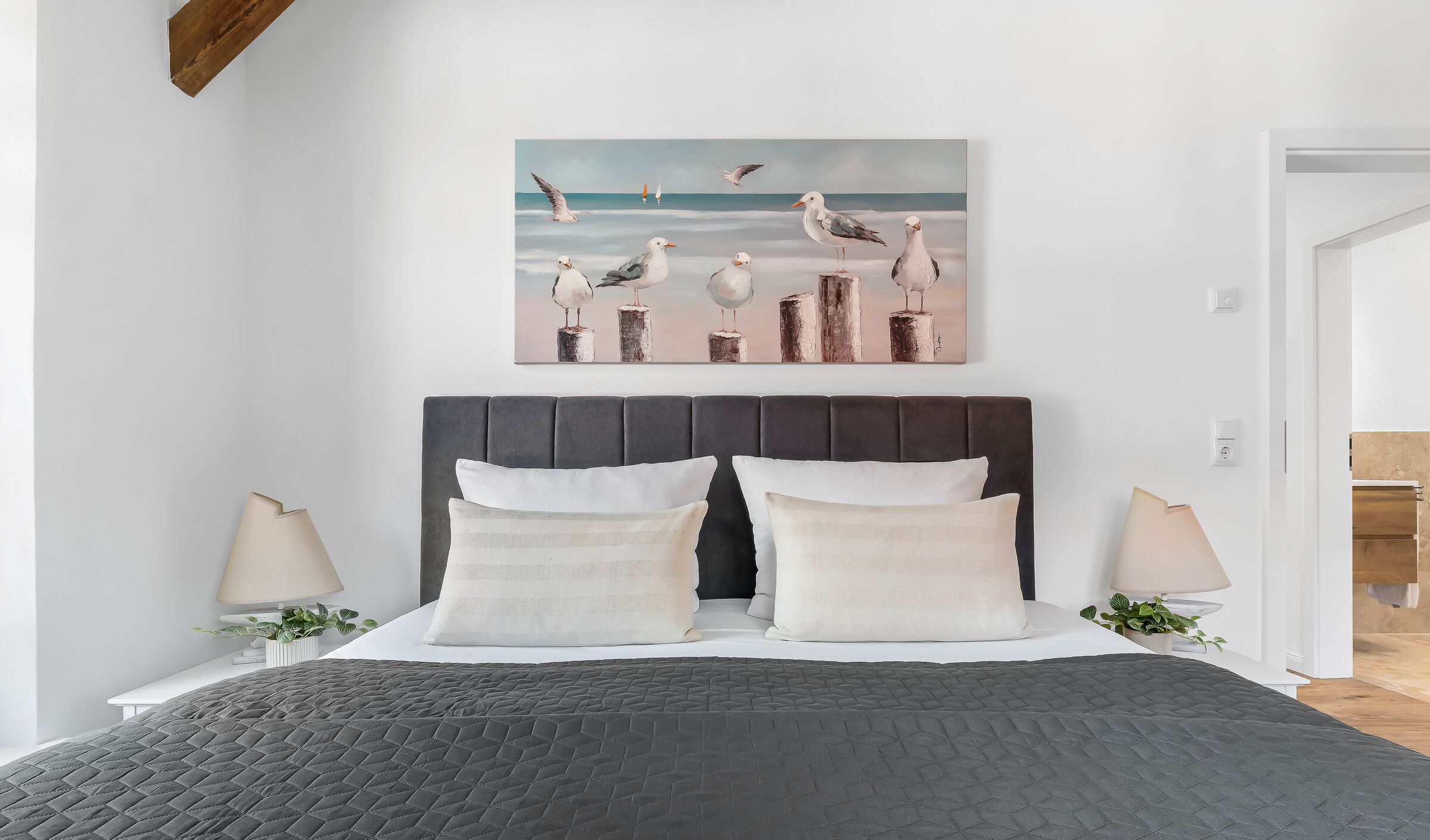 A bedroom with a bed, two bedside lamps, a painting of seagulls, and a view of a wooden door.