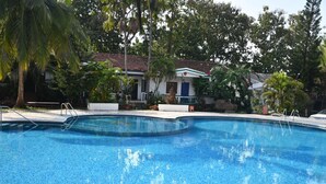 Una piscina con borde de azulejos, rodeada de abundante vegetación y un edificio con tejado rojo al fondo.