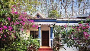 Una casa con una escalera roja que lleva a una puerta de madera, rodeada de vegetación exuberante y flores rosadas.
