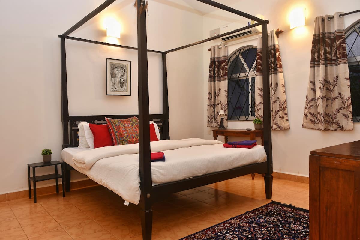 Una cama con dosel con sábanas blancas, una mesita de noche con una planta y una alfombra estampada.