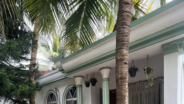 Un edificio con fachada blanca, detalles en verde y un acceso con azulejos. Se observan escalones que conducen a una puerta, una estatua blanca y diferentes plantas y árboles en el entorno.