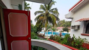 Un balcón con puerta roja, una silla blanca y vista a una piscina y palmeras.