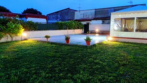 Un patio con plantas en macetas, un edificio blanco con techo rojo y una estructura pequeña con toldo blanco.