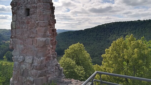 Une tour de pierre surmontée d’une rambarde métallique, entourée d’arbres verdoyants et bordée d’une colline boisée.